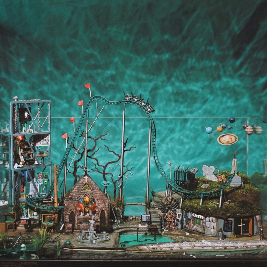 Portada del disco All Inside Aquarium de BODEGA. Fotografía de una maqueta retro hecha a mano dentro de una pecera de agua real, que incluye una mini montaña rusa verde, una iglesia gótica, planetas de canica y un escritorio de oficina central.